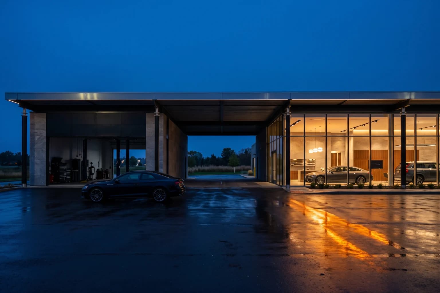 An automotive campus at blue hour, the service bay dark and the showroom lit across a wet forecourt.