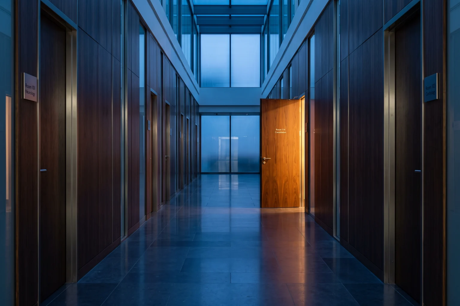 A clinic corridor of identical closed doors with one door open and lit.